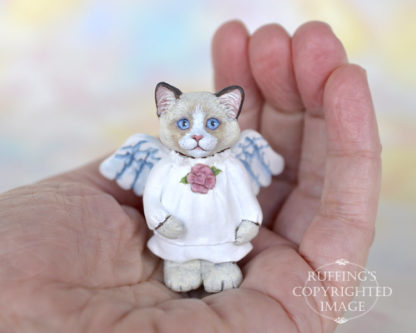 A bi-color ragdoll kitten with angel wings and a rose at her neckline sits in the palm of a hand.