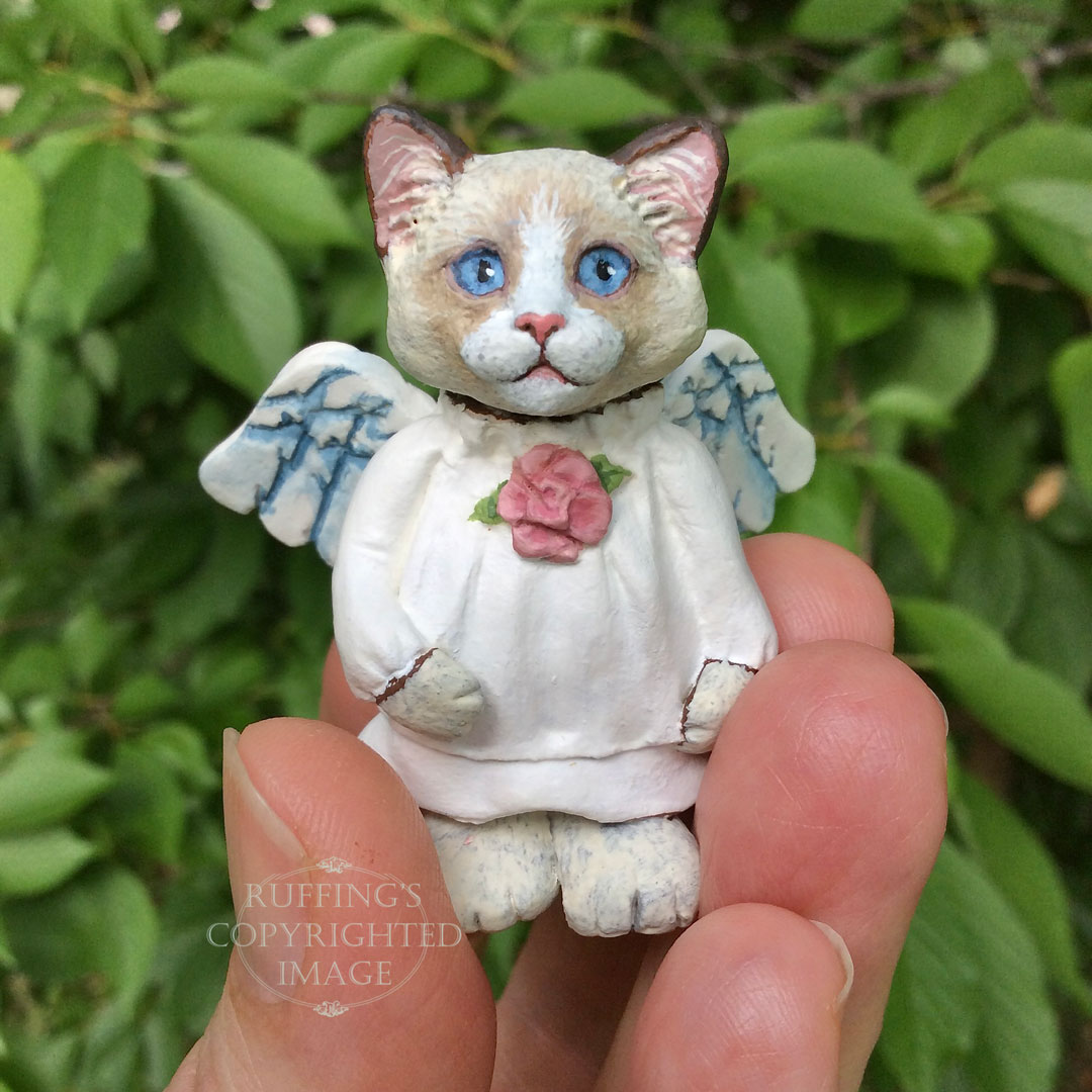 A bi-color ragdoll kitten with angel wings and a rose at her neckline is held between fingertips.