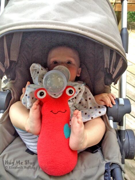 Baby balancing her bottle on her red fleece Hug Me Slug plush toy