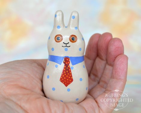 A tan bunny figurine with blue polka dots, a blue collar, and a red tie with yellow polka dots, in the palm of a hand.