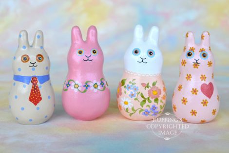 Four bunny figurines, one tan with blue polka dots and a red tie, one mauve-ink with flowers, one peach and white with flowers, and one peach with flowers. 