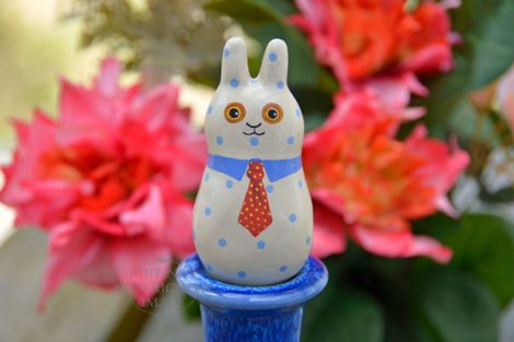 A tan bunny figurine with blue polka dots, a blue collar, and a red tie with yellow polka dots, on a pedestal in front of coral-red flowers. 