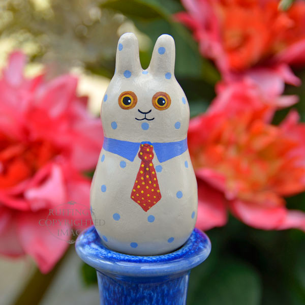 A tan bunny figurine with blue polka dots, a blue collar, and a red tie with yellow polka dots, on a pedestal in front of coral-red flowers.