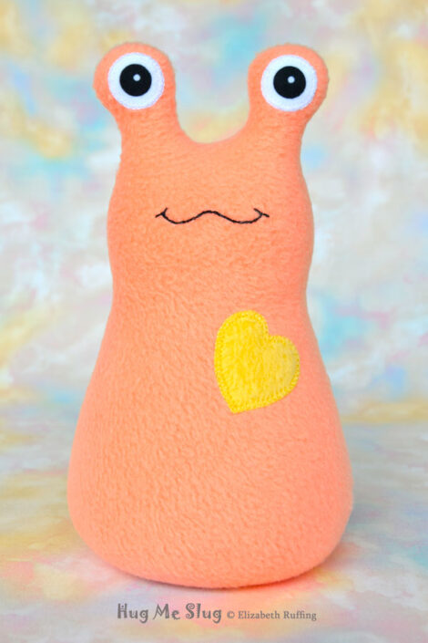 A smiling soft light orange fleece stuffed animal slug with a yellow heart