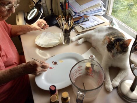 Artist Anne Ruffing painting at her desk with her calico cat sitting nearby