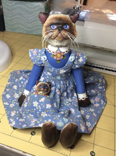A Himalayan cat art doll in a blue floral pinafore sits with a Himalayan kitten in her pocket.