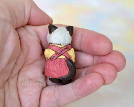 Miniature Himalayan kitten figurine in a mauve pinafore with a painted gold blouse, sitting in a hand, back view
