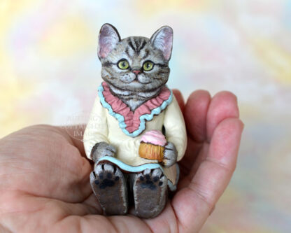 An American Shorthair silver tabby cat figurine with a ruffled mauve collar with pale turquoise trim and a pale cream colored dress sits with a pink frosted cupcake.