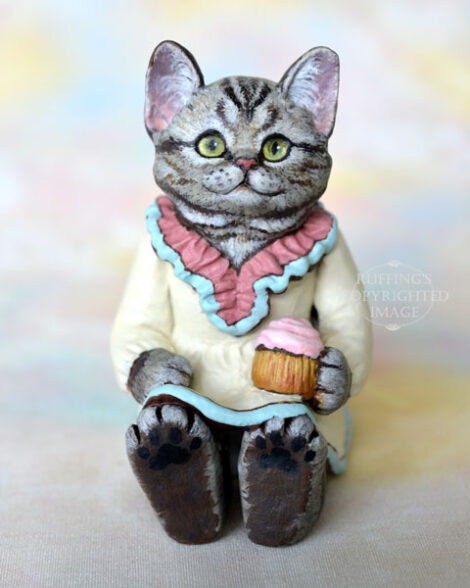 An American Shorthair silver tabby cat figurine with a ruffled mauve collar with pale turquoise trim and a pale cream colored dress sits with a pink frosted cupcake.