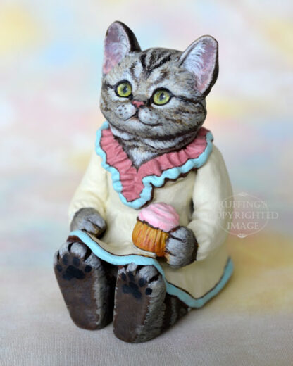 An American Shorthair silver tabby cat figurine with a ruffled mauve collar with pale turquoise trim and a pale cream colored dress sits with a pink frosted cupcake.