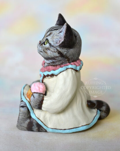 An American Shorthair silver tabby cat figurine with a ruffled mauve collar with pale turquoise trim and a pale cream colored dress sits with a pink frosted cupcake.