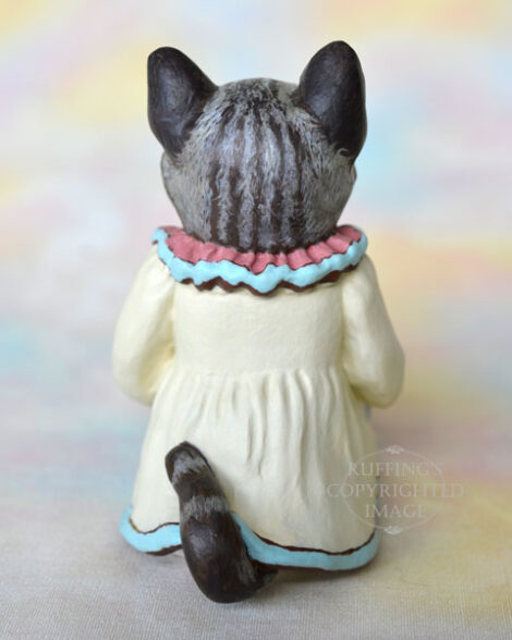 An American Shorthair silver tabby cat figurine with a ruffled mauve collar with pale turquoise trim and a pale cream colored dress sits with a pink frosted cupcake.