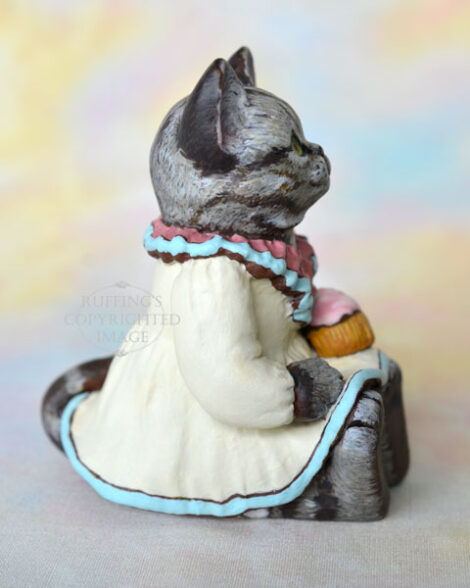 An American Shorthair silver tabby cat figurine with a ruffled mauve collar with pale turquoise trim and a pale cream colored dress sits with a pink frosted cupcake.