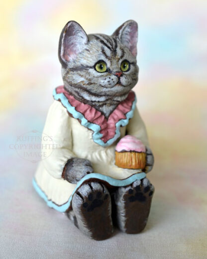 An American Shorthair silver tabby cat figurine with a ruffled mauve collar with pale turquoise trim and a pale cream colored dress sits with a pink frosted cupcake.