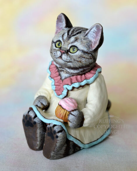 An American Shorthair silver tabby cat figurine with a ruffled mauve collar with pale turquoise trim and a pale cream colored dress sits with a pink frosted cupcake.