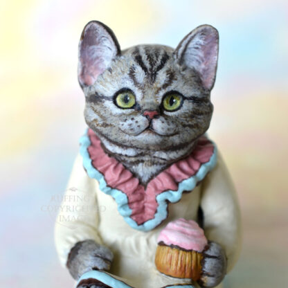 An American Shorthair silver tabby cat figurine with a ruffled mauve collar with pale turquoise trim and a pale cream colored dress sits with a pink frosted cupcake.