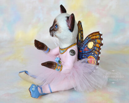 A Siamese cat art doll with bright blue eyes, a lavender-pink tutu, and multicolored wings sits on a soft rainbow colored background.