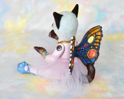 A Siamese cat art doll with bright blue eyes, a lavender-pink tutu, and multicolored wings sits on a soft rainbow colored background.