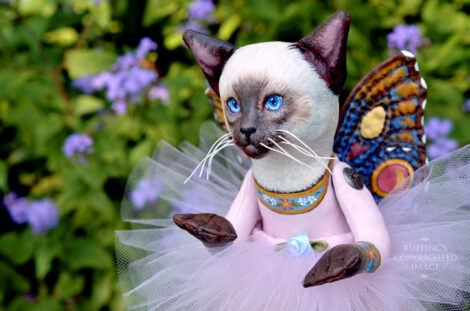 A Siamese cat art doll with bright blue eyes, a lavender-pink tutu, and multicolored wings hovers in front of blue-purple flowers.