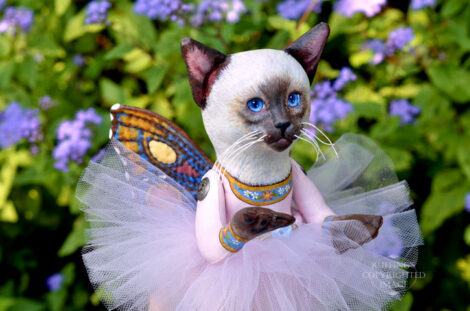 A Siamese cat art doll with bright blue eyes, a lavender-pink tutu, and multicolored wings hovers in front of blue-purple flowers.