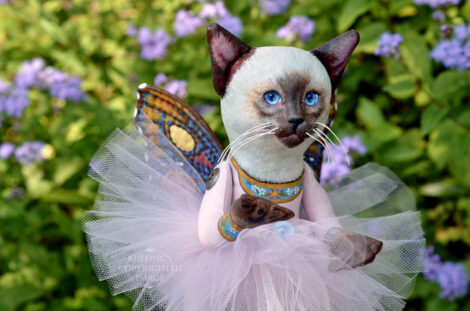 A Siamese cat art doll with bright blue eyes, a lavender-pink tutu, and multicolored wings hovers in front of blue-purple flowers.