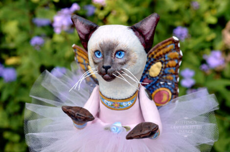 A Siamese cat art doll with bright blue eyes, a lavender-pink tutu, and multicolored wings hovers in front of blue-purple flowers.