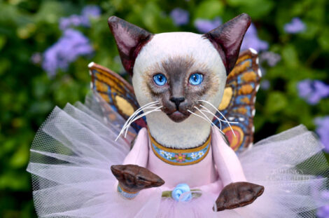 A Siamese cat art doll with bright blue eyes, a lavender-pink tutu, and multicolored wings hovers in front of blue-purple flowers.