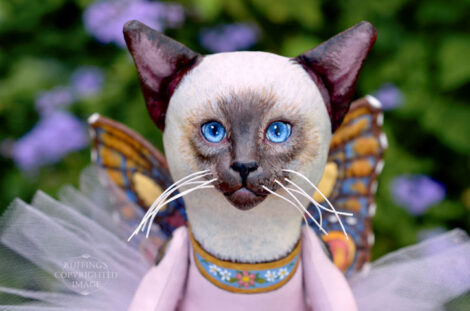 A Siamese cat art doll with bright blue eyes, a lavender-pink tutu, and multicolored wings hovers in front of blue-purple flowers.