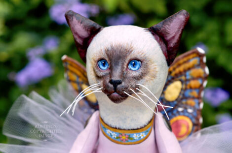 A Siamese cat art doll with bright blue eyes, a lavender-pink tutu, and multicolored wings hovers in front of blue-purple flowers.