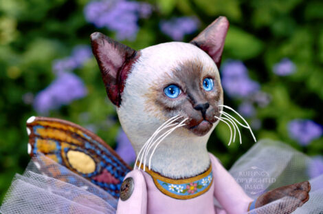 A Siamese cat art doll with bright blue eyes, a lavender-pink tutu, and multicolored wings hovers in front of blue-purple flowers.