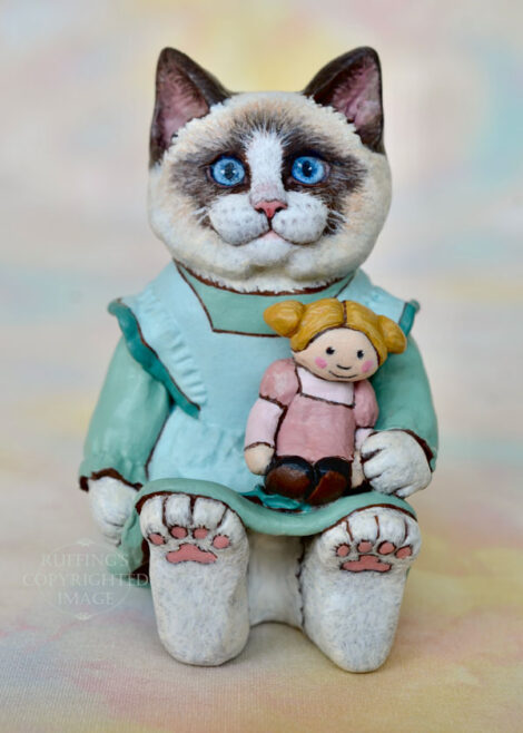 A Ragdoll cat in a light turquoise pinafore holds a blonde ragdoll with ponytails and a mauve dress.