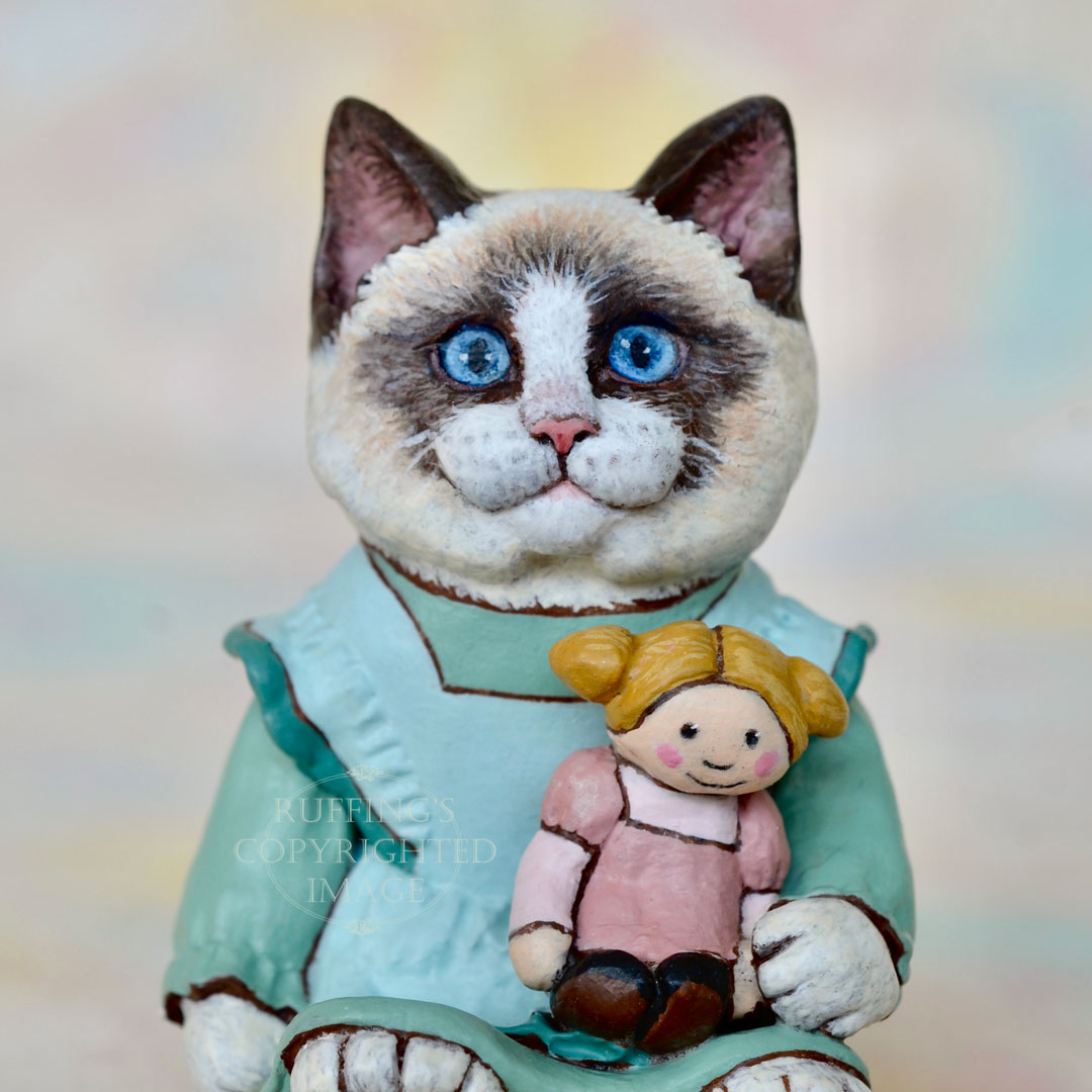 A Ragdoll cat in a light turquoise pinafore holds a blonde ragdoll with ponytails and a mauve dress.