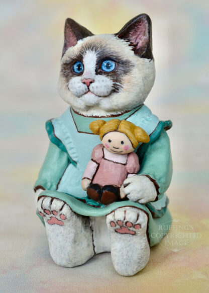 A Ragdoll cat in a light turquoise pinafore holds a blonde ragdoll with ponytails and a mauve dress.