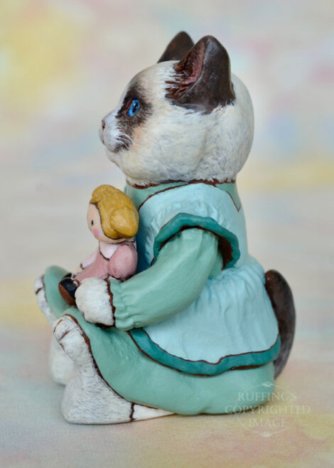 A Ragdoll cat in a light turquoise pinafore holds a blonde ragdoll with ponytails and a mauve dress.