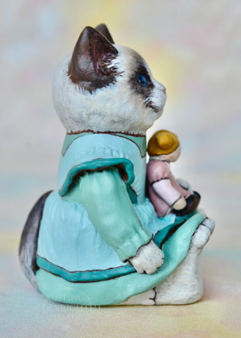 A Ragdoll cat in a light turquoise pinafore holds a blonde ragdoll with ponytails and a mauve dress.