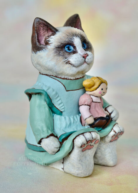 A Ragdoll cat in a light turquoise pinafore holds a blonde ragdoll with ponytails and a mauve dress.