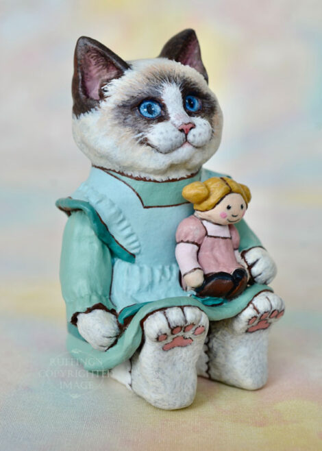 A Ragdoll cat in a light turquoise pinafore holds a blonde ragdoll with ponytails and a mauve dress.