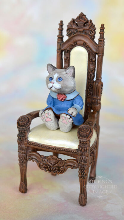 A bi-color ragdoll kitten in a blue dress with a light blue collar and a mauve rose in the middle of her neckline sits in an ornate high-backed chair