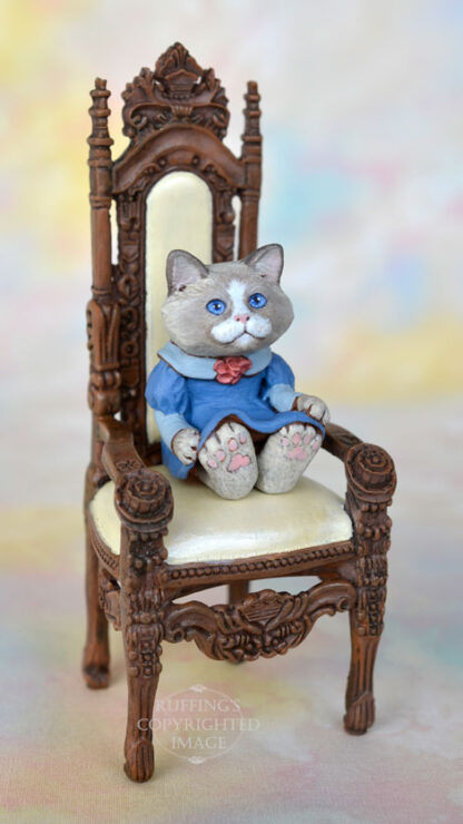 A bi-color ragdoll kitten in a blue dress with a light blue collar and a mauve rose in the middle of her neckline sits in an ornate high-backed chair